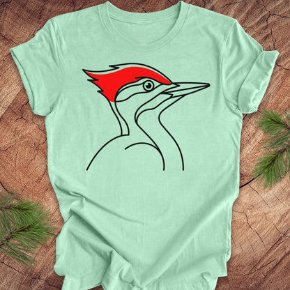 A mint green short-sleeve t-shirt featuring a stylized line art graphic of a woodpecker with a bright red crest, laid flat on a rustic wood background with pine branches in the corners.