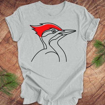 A light gray short-sleeve t-shirt featuring a minimalist line art graphic of a red-breasted woodpecker on the chest, laid flat on a rustic wood background with pine needles in the corners.