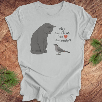 A light gray graphic t-shirt laid flat on a rustic wood background with pine branches. The shirt features a printed illustration of a gray cat looking at a small gray bird, with the text 'why can't we be friends?' and a small red heart.