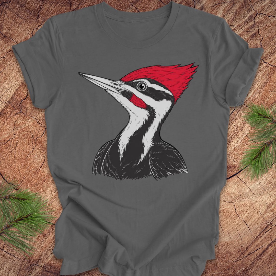 A dark gray short-sleeve t-shirt featuring a graphic print of a Pileated Woodpecker, laid flat on a rustic wood background with pine branches in the corners.