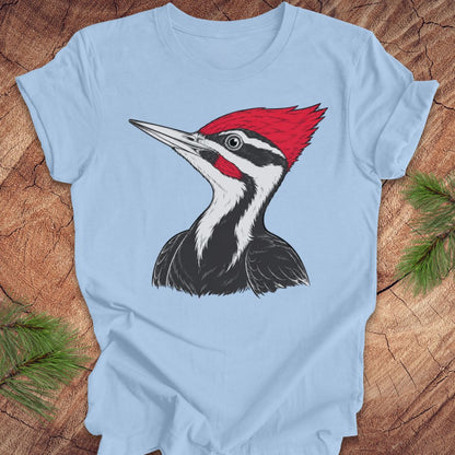 A light blue short-sleeve t-shirt with a graphic of a woodpecker on the front, laid flat on a rustic wood background with pine branches in the corners.