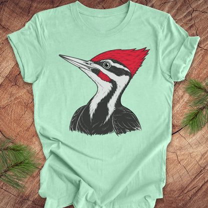 A mint green short-sleeve t-shirt featuring a detailed graphic of a Pileated Woodpecker on the chest, laid flat on a rustic wood background with pine branches in the corners.