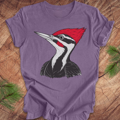 A purple short-sleeve crewneck t-shirt featuring a graphic print of a Pileated Woodpecker, laid flat on a rustic wood background with pine needles in the corners.