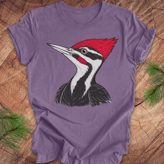 A purple short-sleeve crewneck t-shirt featuring a graphic print of a Pileated Woodpecker, laid flat on a rustic wood background with pine needles in the corners.