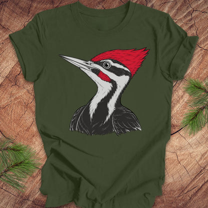 An olive green short-sleeve t-shirt featuring a detailed graphic of a red, white, and black woodpecker on the front, laid flat on a rustic wood background with pine branches.