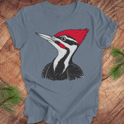 A slate blue short-sleeve t-shirt featuring a detailed graphic of a Pileated Woodpecker on the front, displayed flat on a rustic wood background with pine branches.