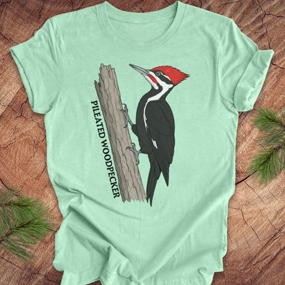 A mint green short-sleeve t-shirt laid flat on a rustic wood surface, featuring a graphic of a pileated woodpecker on a tree trunk with the text 'PILEATED WOODPECKER'.