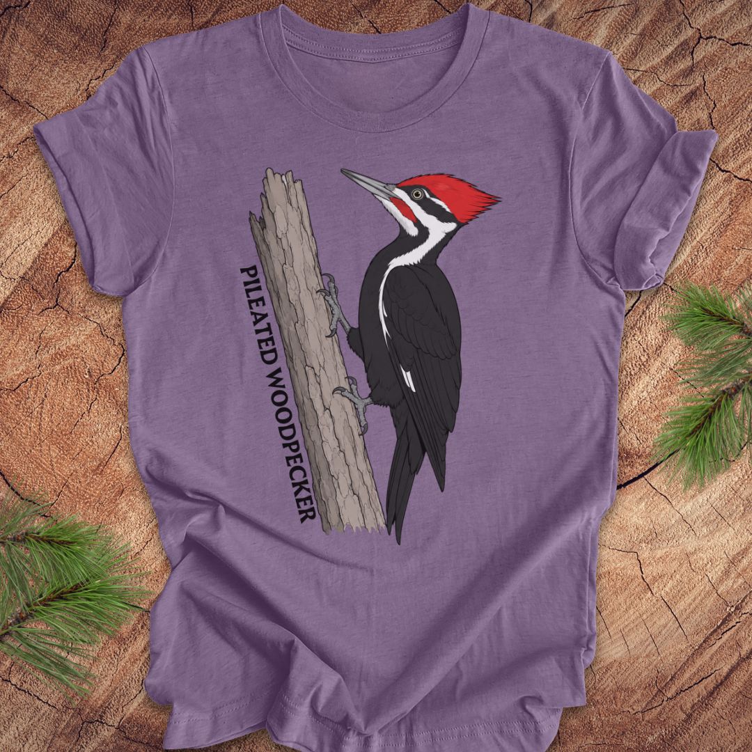 A purple short-sleeve crewneck t-shirt featuring a graphic of a Pileated Woodpecker on a tree trunk with the text 'PILEATED WOODPECKER' printed vertically. The t-shirt is laid flat on a rustic wood background with pine needles in the corners.