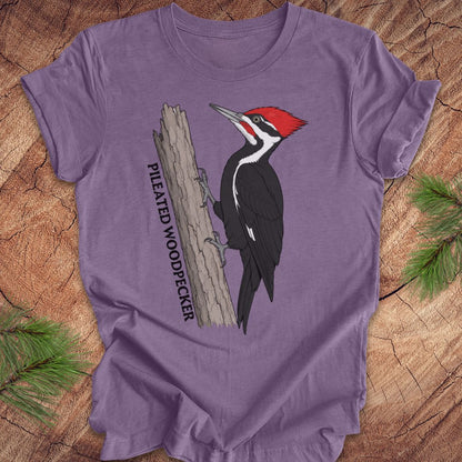 A purple short-sleeve crewneck t-shirt featuring a graphic of a Pileated Woodpecker on a tree trunk with the text 'PILEATED WOODPECKER' printed vertically. The t-shirt is laid flat on a rustic wood background with pine needles in the corners.