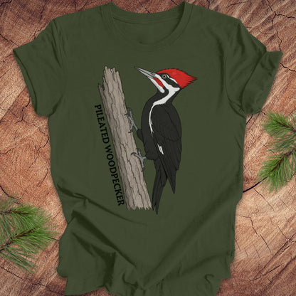 An olive green short-sleeve t-shirt featuring a graphic of a Pileated Woodpecker on a tree trunk with the text 'PILEATED WOODPECKER' printed vertically. The t-shirt is laid flat on a rustic wood background with pine branches in the corners.