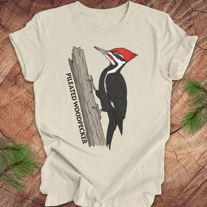 A cream-colored crewneck t-shirt featuring a graphic of a pileated woodpecker on a tree trunk with the text 'PILEATED WOODPECKER' printed vertically. The t-shirt is laid flat on a rustic wood background with pine branches in the corners.