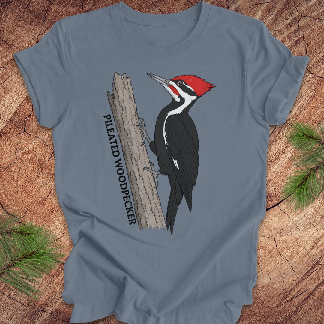 A slate blue crewneck t-shirt featuring a graphic of a Pileated Woodpecker on a tree trunk with the text 'PILEATED WOODPECKER' printed vertically. The t-shirt is displayed flat on a rustic wood background with pine needles in the corners.
