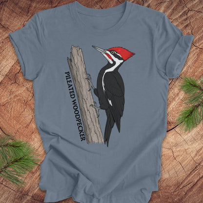 A slate blue crewneck t-shirt featuring a graphic of a Pileated Woodpecker on a tree trunk with the text 'PILEATED WOODPECKER' printed vertically. The t-shirt is displayed flat on a rustic wood background with pine needles in the corners.