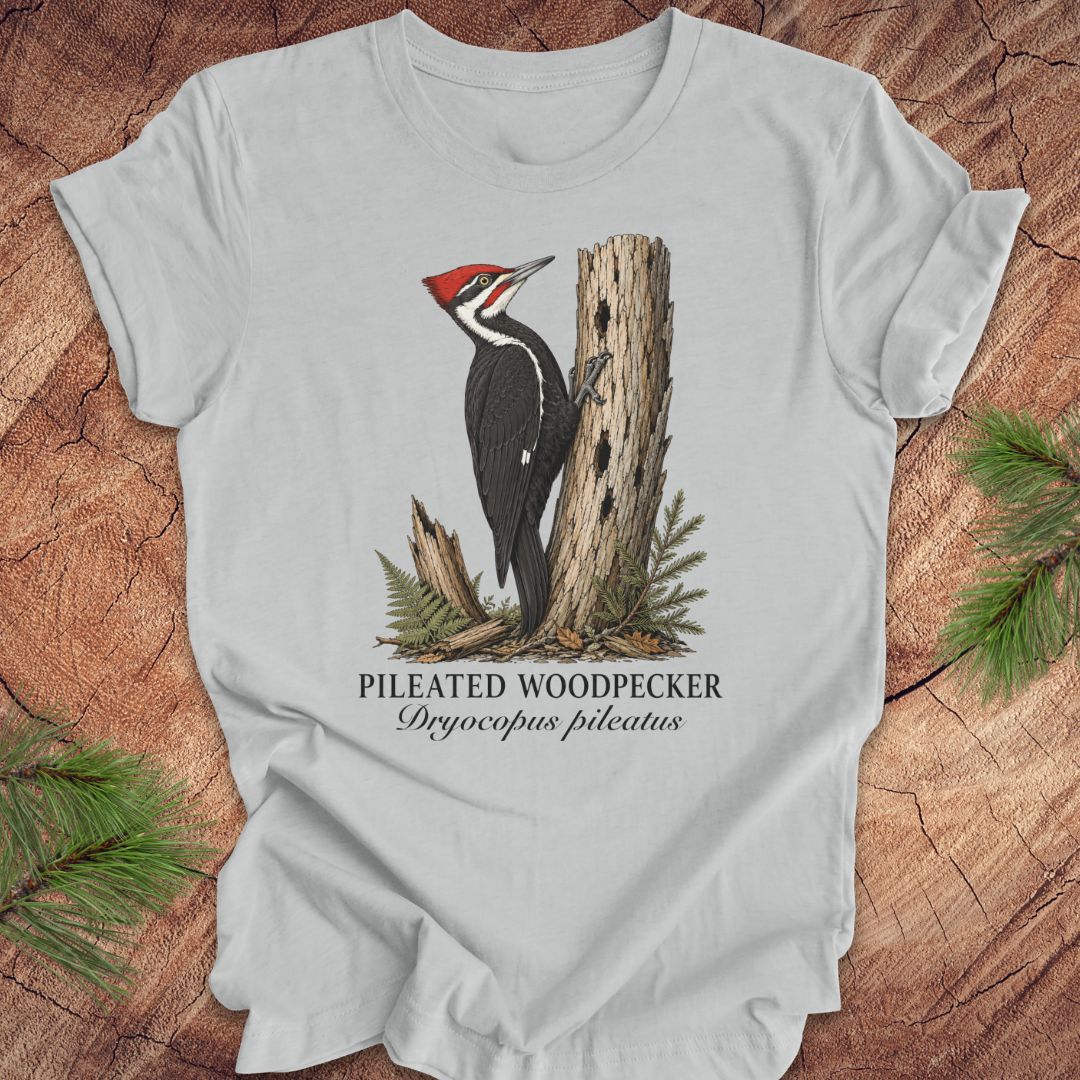 Gray t-shirt with a pileated woodpecker graphic and text on a wooden surface with pine branches.
