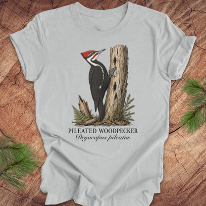Gray t-shirt with a pileated woodpecker graphic and text on a wooden surface with pine branches.