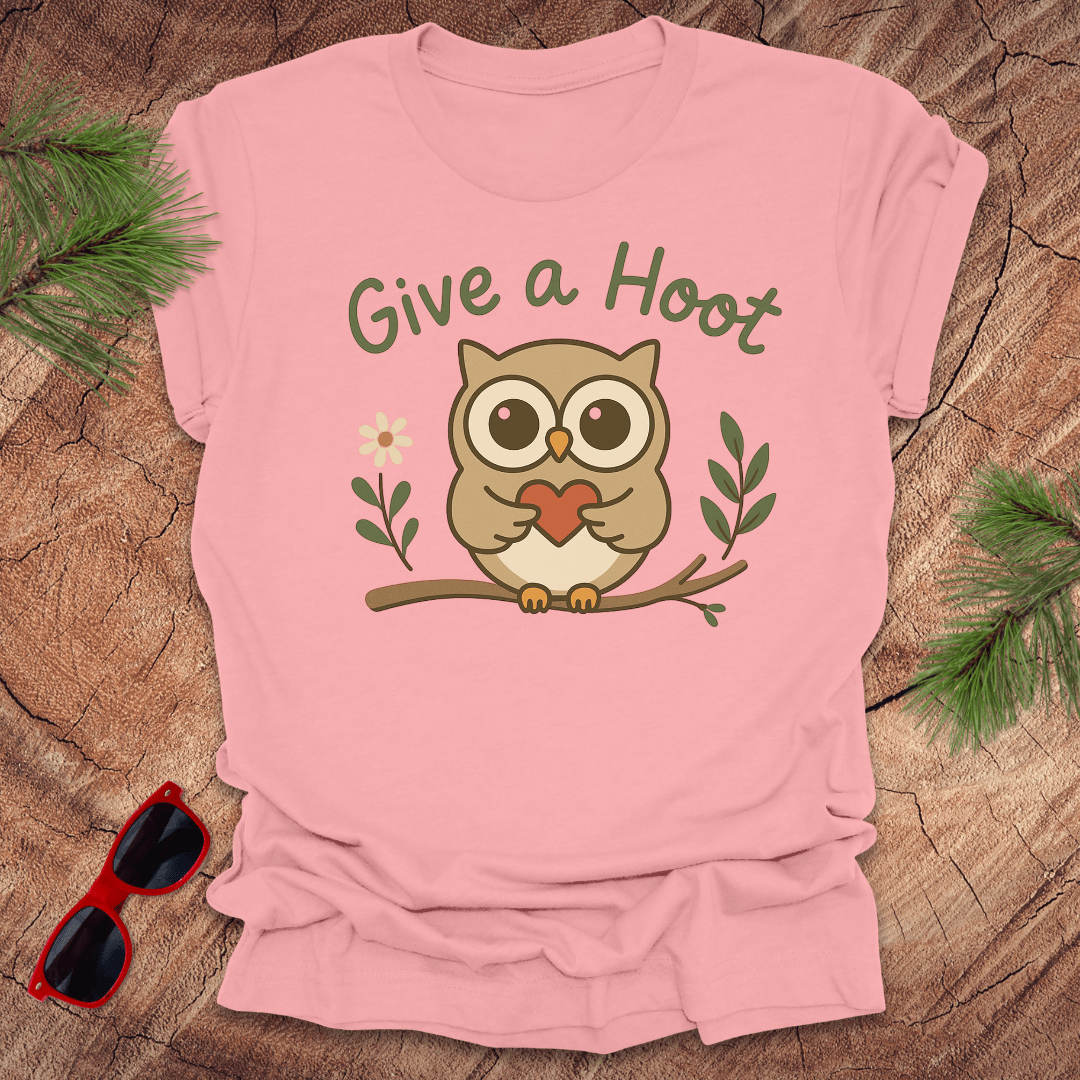 Pink t-shirt with owl graphic and 'Give a Hoot' text on a wooden surface with sunglasses and greenery.