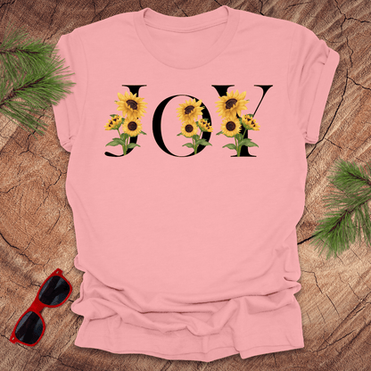 Pink t-shirt with 'JOY' and sunflower design on a wooden surface with sunglasses and greenery.
