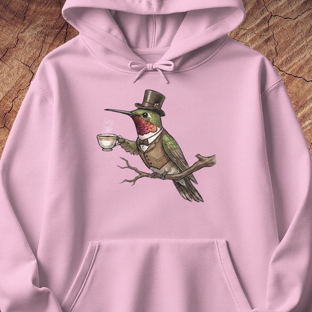 A light pink hoodie laid flat on a textured wood background, featuring a central graphic of a green bird wearing a brown top hat and vest while holding a teacup.