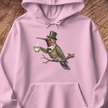 A light pink hoodie laid flat on a textured wood background, featuring a central graphic of a green bird wearing a brown top hat and vest while holding a teacup.