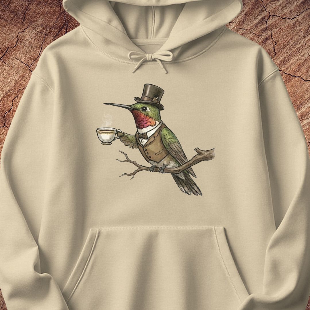 A cream-colored hoodie featuring a detailed graphic of a hummingbird dressed in a top hat, waistcoat, and bow tie, holding a teacup on a branch. The hoodie is displayed flat against a textured brown wood background.