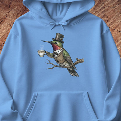 A light blue pullover hoodie laid flat on a rustic wood background, featuring a central graphic of a hummingbird dressed in a top hat and vest while holding a teacup.