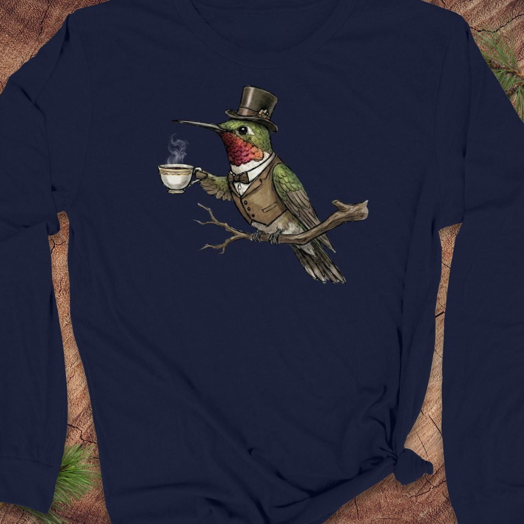 A navy blue long-sleeve t-shirt featuring a graphic print of a green hummingbird dressed in a top hat, waistcoat, and bow tie while holding a steaming teacup. The shirt is styled flat on a rustic wood background with pine needles.