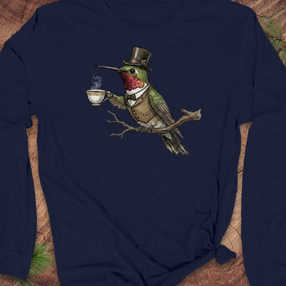 A navy blue long-sleeve t-shirt featuring a graphic print of a green hummingbird dressed in a top hat, waistcoat, and bow tie while holding a steaming teacup. The shirt is styled flat on a rustic wood background with pine needles.