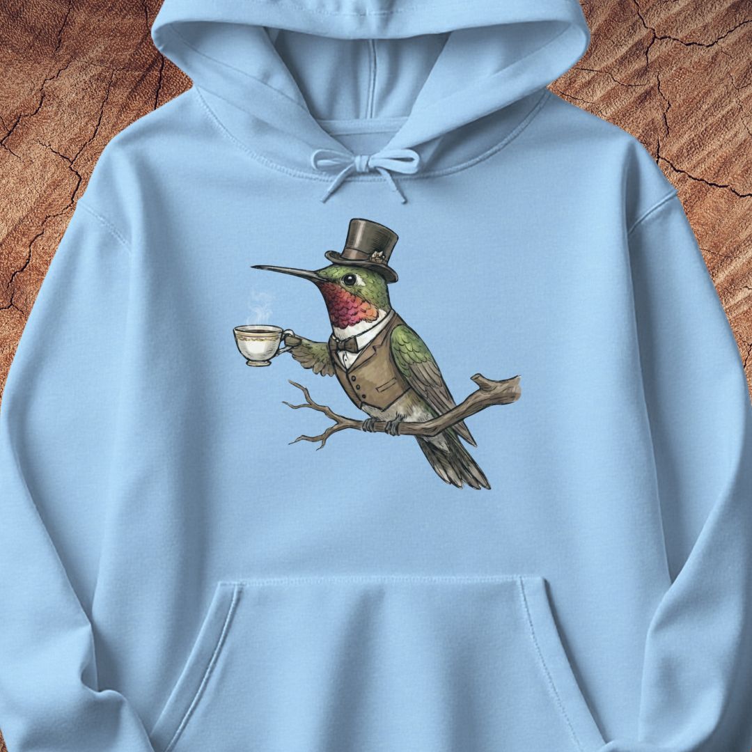 A light blue pullover hoodie featuring a detailed graphic of a green hummingbird dressed in a brown top hat, vest, and bow tie while perched on a branch and holding a teacup. The hoodie is displayed flat against a textured brown wood background.