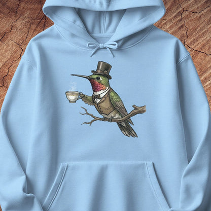 A light blue pullover hoodie featuring a detailed graphic of a green hummingbird dressed in a brown top hat, vest, and bow tie while perched on a branch and holding a teacup. The hoodie is displayed flat against a textured brown wood background.
