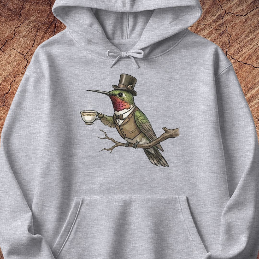 A light gray heathered hoodie featuring a whimsical graphic of a green hummingbird dressed in a brown top hat and vest, perched on a branch and holding a teacup. The hoodie is displayed flat against a rustic wood background.