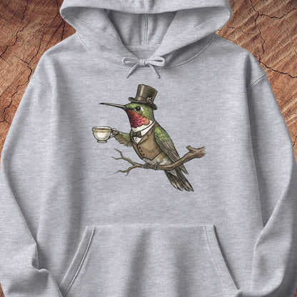 A light gray heathered hoodie featuring a whimsical graphic of a green hummingbird dressed in a brown top hat and vest, perched on a branch and holding a teacup. The hoodie is displayed flat against a rustic wood background.