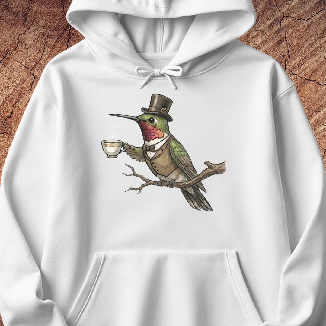 A white pullover hoodie featuring a detailed graphic of a green hummingbird wearing a brown top hat and vest, perched on a branch and holding a teacup. The hoodie is displayed flat against a textured brown wood background.