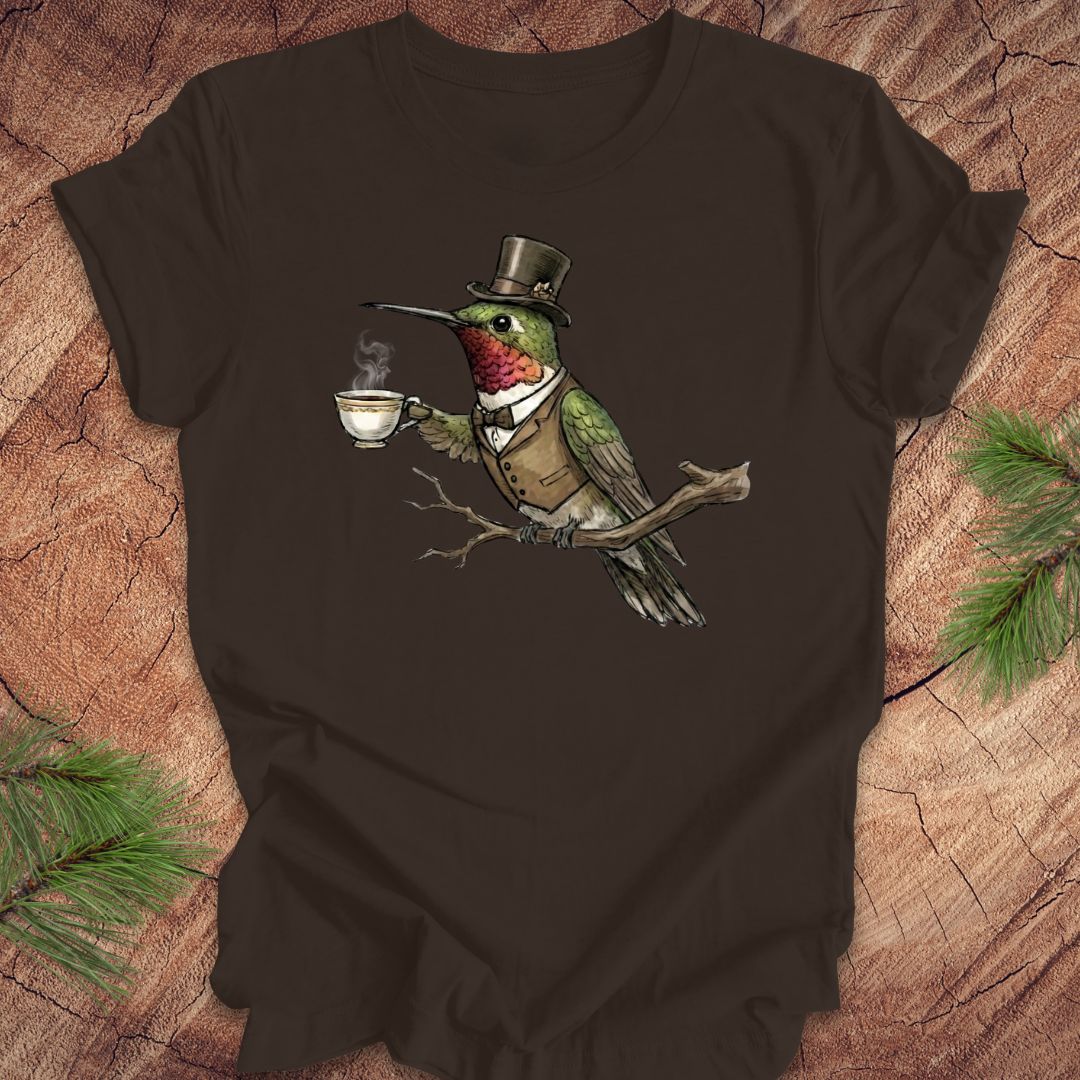 Brown t-shirt with a graphic of a hummingbird in a top hat and monocle holding a cup on a wooden surface.