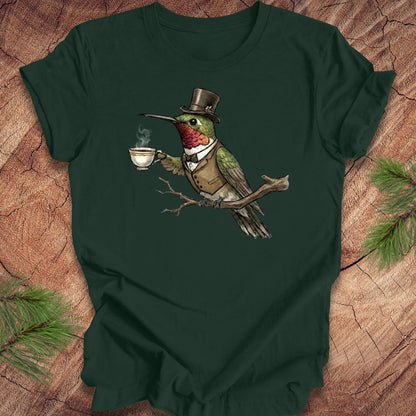 Dark green t-shirt with a cartoon hummingbird wearing a top hat and holding a cup on a wooden surface.