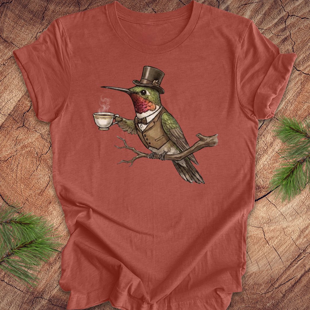 Clay colored t-shirt with a cartoon hummingbird wearing a top hat and holding a cup on a wooden surface.