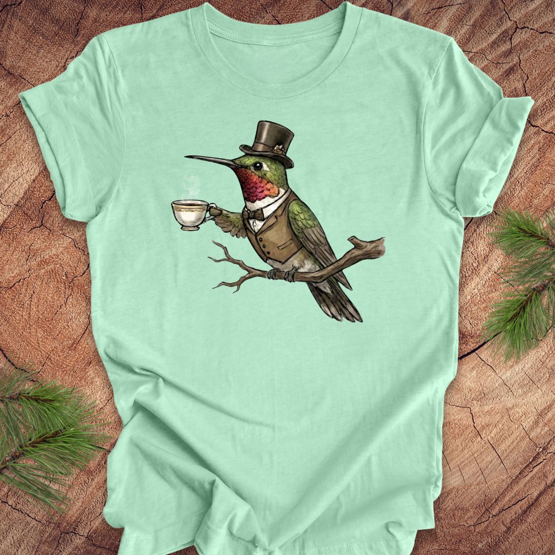 Mint green t-shirt with a cartoon hummingbird wearing a top hat and holding a cup on a wooden surface.