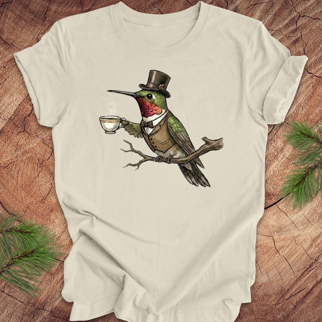 Beige t-shirt with a hummingbird wearing a top hat and holding a cup design on a wooden surface.