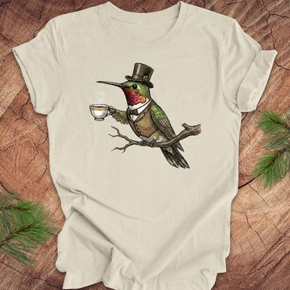 Beige t-shirt with a hummingbird wearing a top hat and holding a cup design on a wooden surface.
