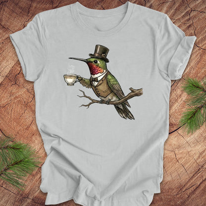 Gray t-shirt with a cartoon hummingbird wearing a top hat and holding a cup on a wooden surface.