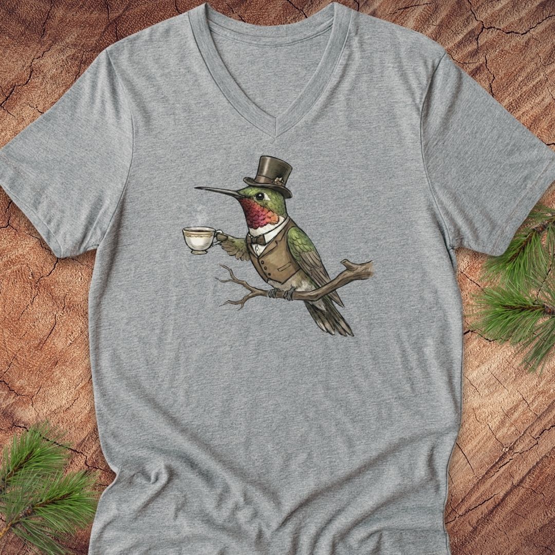 A heather gray V-neck t-shirt featuring a graphic of a green hummingbird wearing a top hat and waistcoat while holding a teacup, laid flat on a rustic wood background with pine needles.
