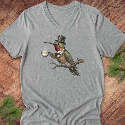 A heather gray V-neck t-shirt featuring a graphic of a green hummingbird wearing a top hat and waistcoat while holding a teacup, laid flat on a rustic wood background with pine needles.