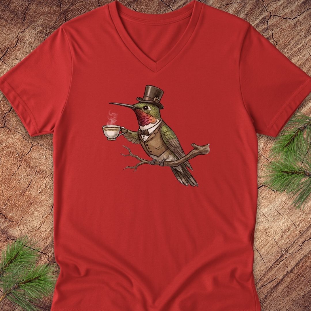 A red V-neck t-shirt laid flat on a rustic wood background with pine branches in the corners. The shirt features a central graphic of a green bird wearing a brown top hat and vest while holding a white teacup.