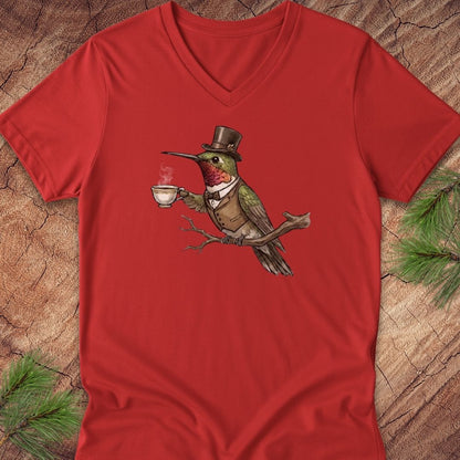 A red V-neck t-shirt laid flat on a rustic wood background with pine branches in the corners. The shirt features a central graphic of a green bird wearing a brown top hat and vest while holding a white teacup.