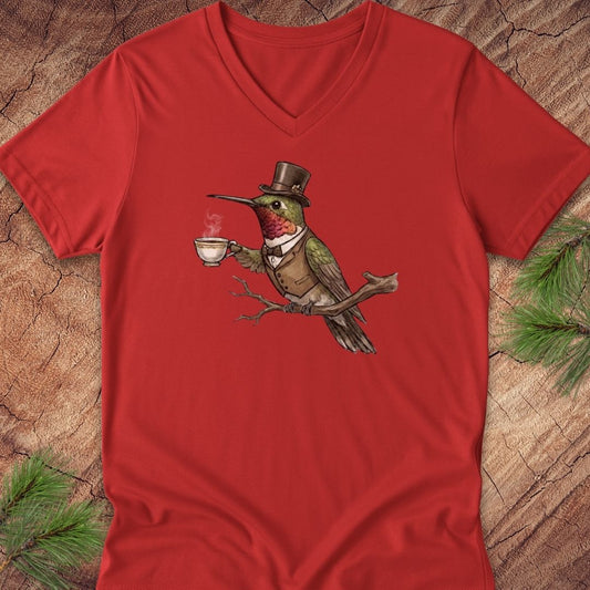 A red V-neck t-shirt laid flat on a rustic wood background with pine branches in the corners. The shirt features a central graphic of a green bird wearing a brown top hat and vest while holding a white teacup.