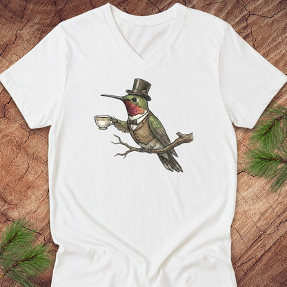 A white v-neck t-shirt featuring a graphic of a hummingbird dressed in a top hat and vest, holding a teacup on a branch. The shirt is displayed flat on a rustic wood background with pine needles in the corners.