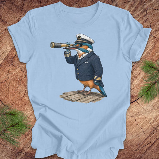 Light blue t-shirt with a kingfisher bird in a captain's outfit and spyglass on a wooden surface.