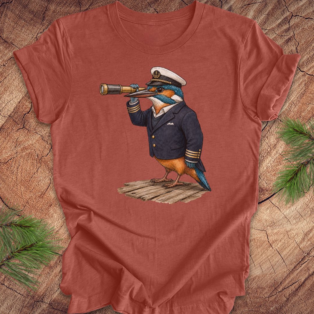 Clay colored t-shirt with a kingfisher bird in a captain's outfit and spyglass on a wooden surface.