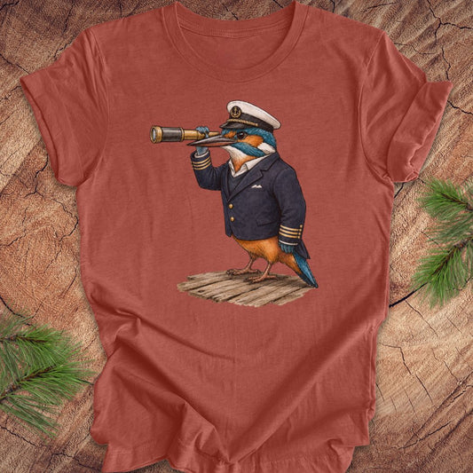 Clay colored t-shirt with a kingfisher bird in a captain's outfit and spyglass on a wooden surface.