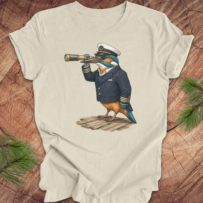 Beige t-shirt with a kingfisher bird in a captain's outfit and spyglass on a wooden surface.