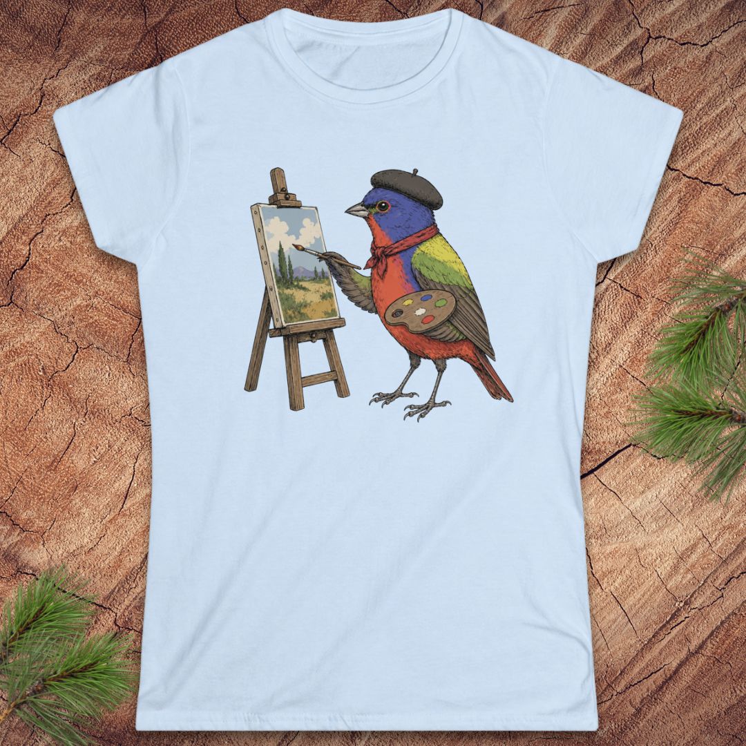 A light blue short-sleeve crewneck t-shirt featuring a colorful graphic of a bird painting on an easel, laid flat on a rustic wood background with pine needles.